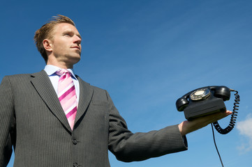 Naklejka premium Businessman standing waiting outdoors holding old fashioned black rotary dial telephone in blue sky copy space