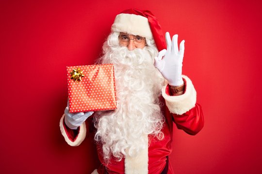 Middle age man wearing Santa Claus costume holding gift over isolated red background doing ok sign with fingers, excellent symbol