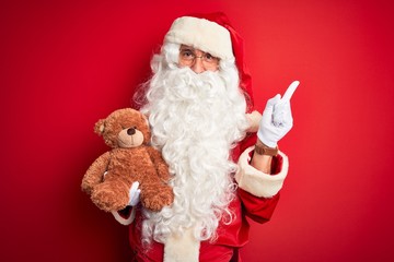 Middle age man wearing Santa Claus costume holding teddy bear over isolated red background very happy pointing with hand and finger to the side