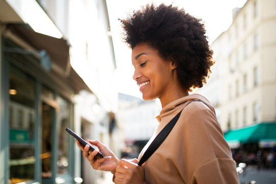 Side portrait of smiling young black woman looking at mobile phone in city