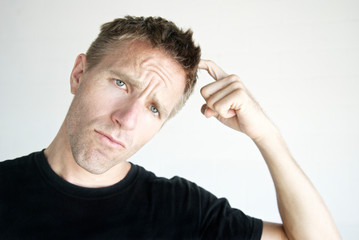 Casual young man in black T-shirt furrowing his brow and scratching his head looking at the camera with a puzzled expression