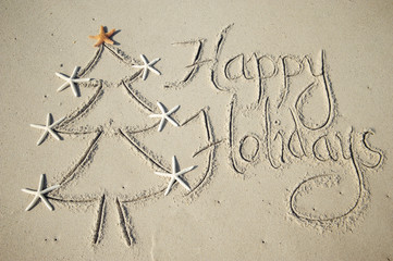 Happy Holidays message handwritten in smooth sand with simple eco-friendly Christmas tree decorated with starfish on the beach