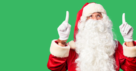 Middle age handsome man wearing Santa Claus costume and beard standing smiling amazed and surprised and pointing up with fingers and raised arms.