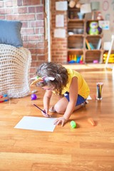 Beautiful toddler wearing glasses and unicorn diadem sitting on the floor drawing using paper and pencils at kindergarten