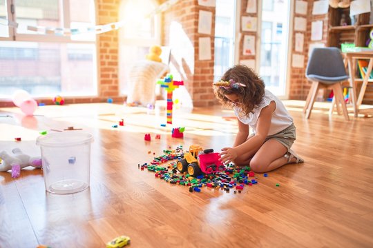 Beautiful toddler wearing glasses and unicorn diadem playing with tractor and building blocks at kindergarten