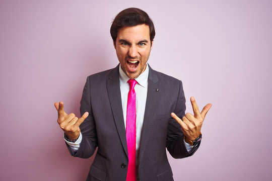 Young handsome businessman wearing suit and tie standing over isolated pink background shouting with crazy expression doing rock symbol with hands up. Music star. Heavy concept.