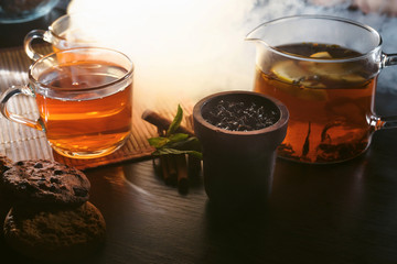 Cafe or tearoom hookah lounge concept. Two cups of aroma herbal black or red tea with some fresh cookies with spices closeup on wooden table in clouds of smoke.
