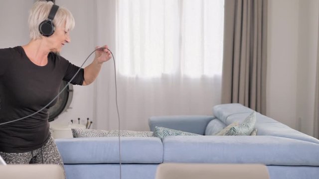 happy mature elderly woman with headphones listen to music and dancing while vacuuming in the living room