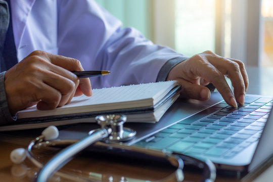 Female doctor hand writing on empty white notebook while working on laptop computer in medical room at clinic or hospital.