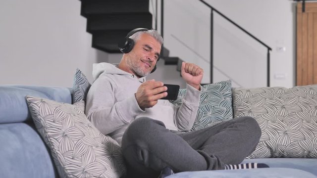 Happy mature man with headphones listen to music wireless and watching video on the sofa holding smartphone