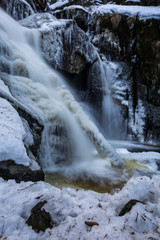 High waterfall in the forest. Water is falling on rock surface. Ice on the long tree trunk. Typical scene in winter period.