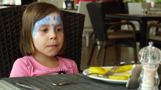 A girl with a painted face is sitting in the dining room. A child with blond hair, 5-6 years old, eats chips and looks into the smartphone.
