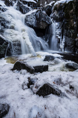 High waterfall in the forest. Water is falling on rock surface. Ice on the long tree trunk. Typical scene in winter period.