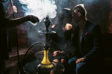 Hookah lounge concept. Beautiful couple woman and man in backlight in love smoking and drinks cocktails in the bar. Hands of hookahman with forceps and kaloud in foreground in clouds of smoke