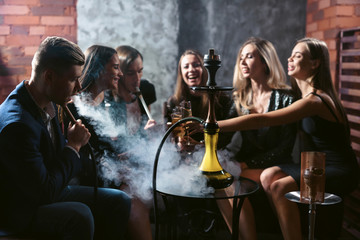 Friends party in hookah lounge. Group of people women and men smoking shisha in cafe or bar, making smoke clouds, having fun, smiling. Relax concept. Friendship - obrazy, fototapety, plakaty
