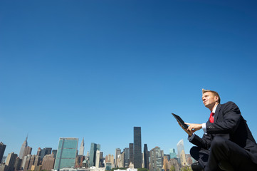 Fototapeta na wymiar Businessman sitting outdoors using a tablet computer in front of the city skyline under clear blue sky copy space