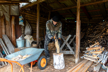 An elderly Russian man hunched wearily chopping wood with an axe in a barn in the village.
