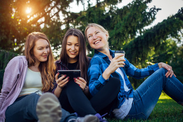 Fototapeta premium Happy young girls have fun laughing sitting on green grass in the park. Students in between lectures. Blonde, brunette and red-hair girl communicate, look at smartphone, concept of female friendship.