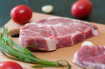 Raw juicy pork steaks with red tomatoes, rosemary and garlic on a cutting board.
