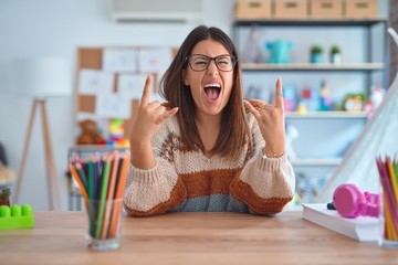 Young beautiful teacher woman wearing sweater and glasses sitting on desk at kindergarten shouting with crazy expression doing rock symbol with hands up. Music star. Heavy music concept. - Powered by Adobe