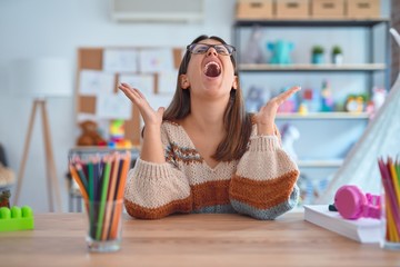Young beautiful teacher woman wearing sweater and glasses sitting on desk at kindergarten crazy and mad shouting and yelling with aggressive expression and arms raised. Frustration concept.