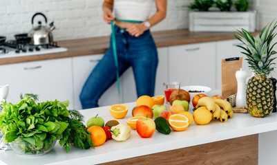 Young slim girl cropped photo with measuring tape standing in the modern kitchen. Fruits and vegetables on the table. Fitness woman and healthy food concept.