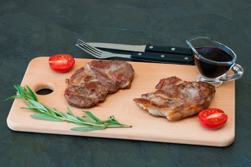 Grilled meat steaks of pork, veal or beef, with rosemary, soy sauce and tomato on cutting board, fork and knife