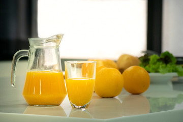 Glass and pitcher of orange juice on white table in kitchen with oranges, bun and salad for breakfast , focus at orange juice.