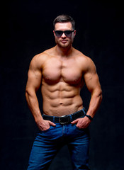 Fototapeta na wymiar A portrait of a hot handsome strong guy man without a shirt against dark background. hands in pockets wearing glasses. Studio photo. Male beauty concept. Closeup.