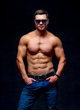 A portrait of a hot handsome strong guy man without a shirt against dark background. hands in pockets wearing glasses. Studio photo. Male beauty concept. Closeup.