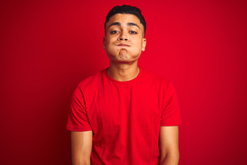 Young brazilian man wearing t-shirt standing over isolated red background puffing cheeks with funny face. Mouth inflated with air, crazy expression.