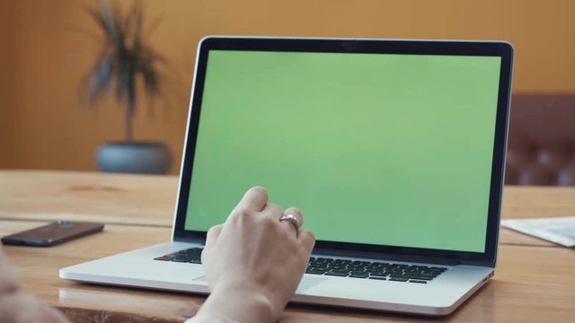 Young woman in pink shirt sitting at the large wooden table and typing on her laptop with chroma key green screen. Stock footage. Chroma key screen for placement of your own content.