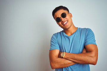 Young brazilian man wearing blue t-shirt and sunglasses over isolated white background happy face smiling with crossed arms looking at the camera. Positive person.