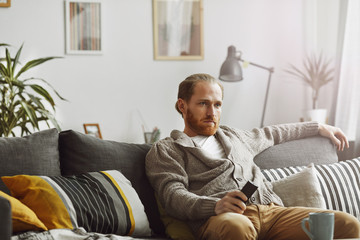 Portrait of bored bearded man watching TV at home and holding remote control while relaxing on...