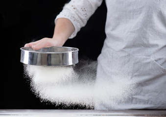 Cook sifts flour through a sieve on a wooden table. Isolated on dark background