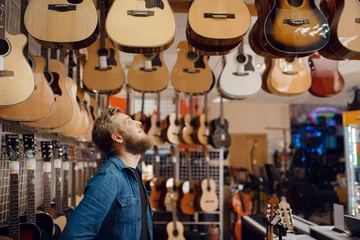 Foto op Plexiglas Muziekwinkel Jonge kerel die akoestische gitaar kiest in muziekwinkel