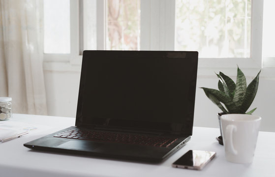 Black laptop is placed on a white desk with in the office with windows and ornamental plants. Business objects concept