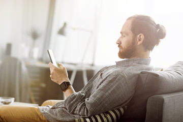 Side view portrait of contemporary bearded man using digital tablet while relaxing on comfortable couch at home, copy space