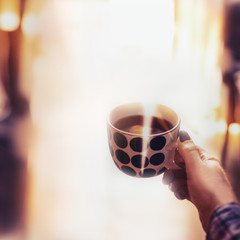 Female hand holding polka dot cup with steamed hot drink at cozy home background with sunlight and bokeh