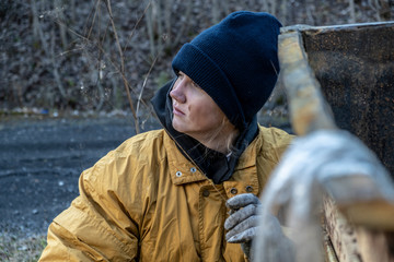 homeless woman in a yellow old ragged jacket and blue hat is sits near the garbage can. poverty.