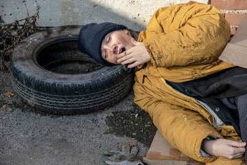 A homeless woman in a yellow old torn jacket and a blue hat lies and sleeping on cardboard on the sidewalk