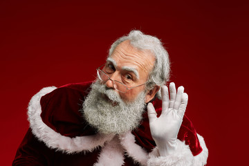 Portrait of classic Santa Claus putting hand to ear trying to hear secrets while standing against red background in studio, copy space