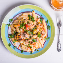 Italian cuisine - penne rigate pasta with sweet pepper sauce with tomatoes, tuna with olives, sprinkled with basil leaves on a plate on a light wooden table, top view