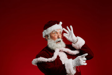 Waist up portrait of surprised Santa Claus pointing to side and adjusting glasses while posing against red background in studio, copy space