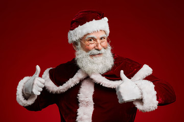 Waist up portrait of cheerful Santa Claus showing thumbs up while standing against red background in studio, copy space