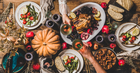 Thanksgiving party table setting. Flat-lay of couple clinking glasses and celebrating at table with roasted chicken, vegetables, fig pie, fruit, candles over wooden table background, top view