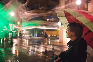 Young girl with short hair and dressed in black, walks through the city at night and raining. Take an umbrella and look at shop windows.