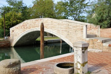 Old stone bridge above a canal, and old stone fountain on the island Torcello. It is located next to the island Burano. Lagoon of Venice. Italy, South Europe.
