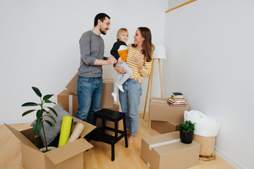Fototapeta na wymiar Young family moving to a new home, standing in empty room next to pile of boxes