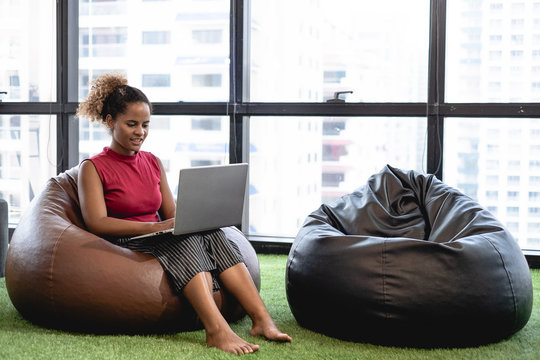 Happy Woman Sitting On Bean Bag In Modern Office Working With Laptop.